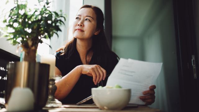 a woman seated at a window in a modern kitchen is reading paperwork and thinking with a bowl of food and a plant in the foreground