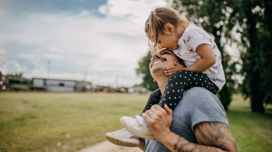 father with tee shirt and tattoos is carrying his young daughter on his shoulders in a park 