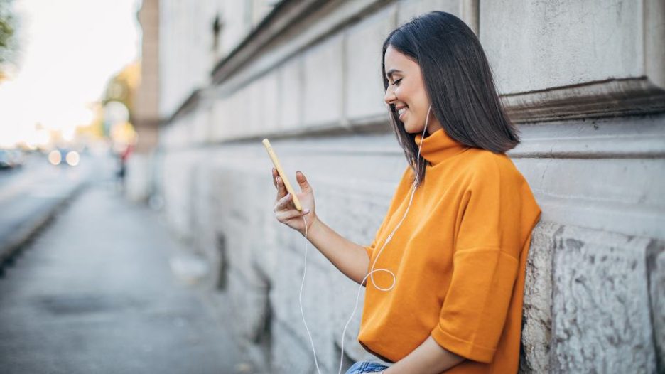smiling woman in an orange jumper is on her phone while resting against a stone wall