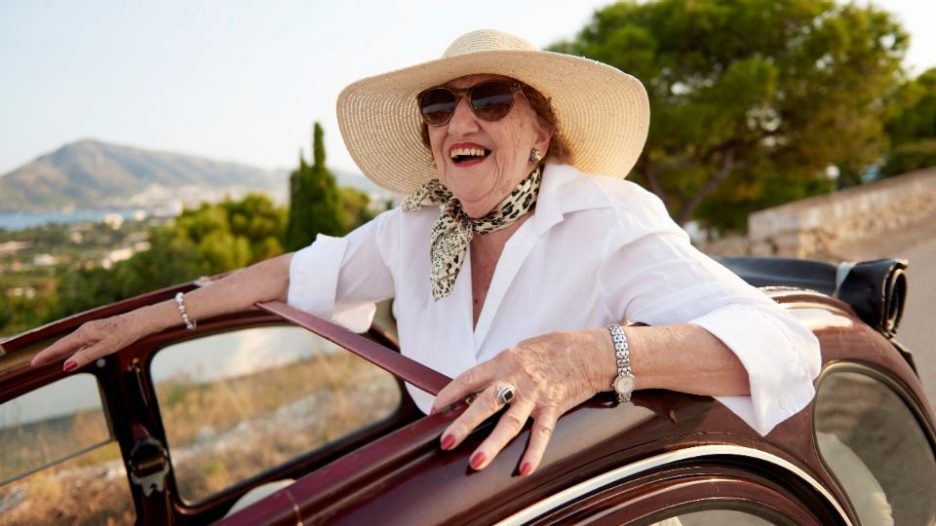 A glamourous elderly woman sitting in a car with a sun hat and sunglasses on