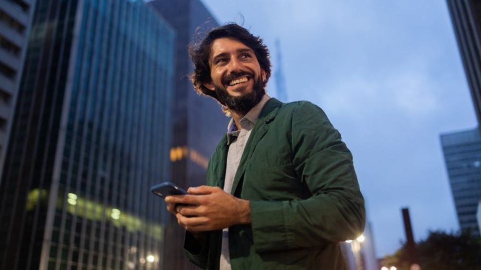 Happy smiling man using his phone while crossing the street of a business district at dusk