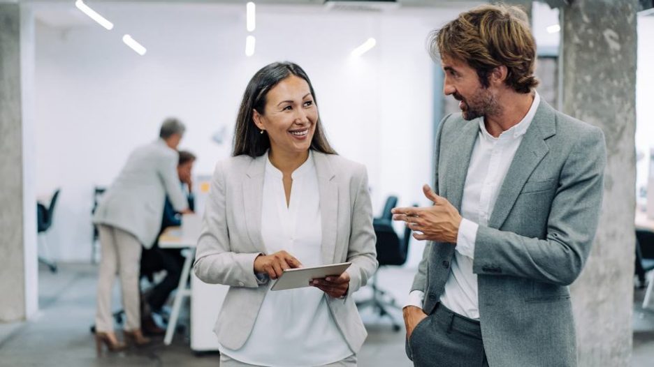 A happy professional woman is chatting with her male coworker whle a third woman looks on