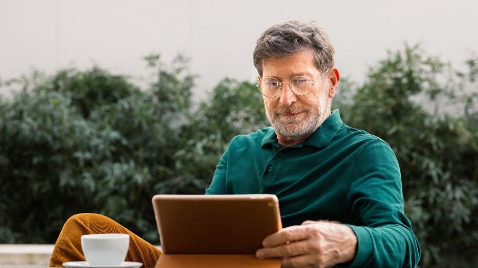 senior man sitting outdoors on terrace, drinking coffee and using digital tablet