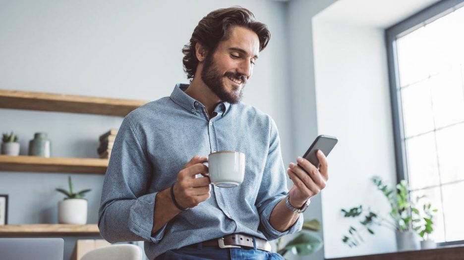 casually dressed man in a modern office is smiling and drinking coffee while looking at his phone