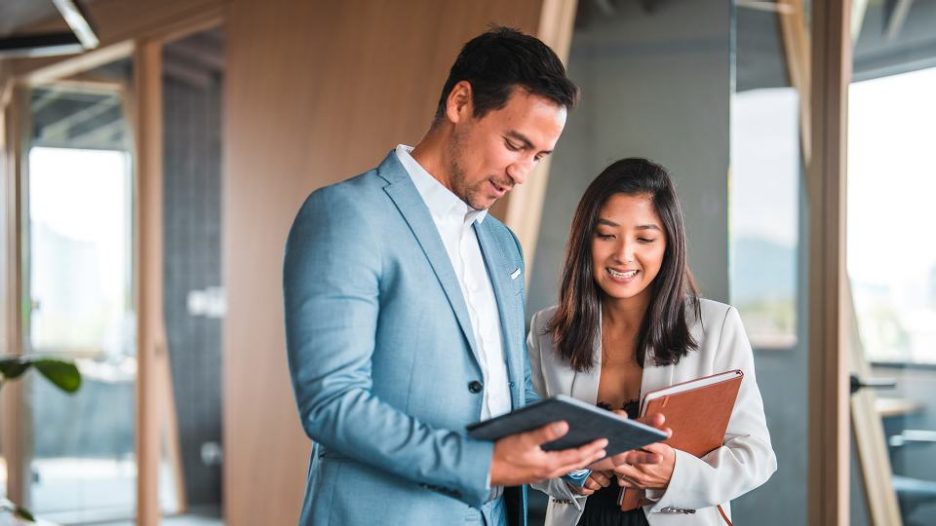 Waist up shot of beautiful woman and asian male boss smiling while working together. They discuss business while looking at a digital tablet.