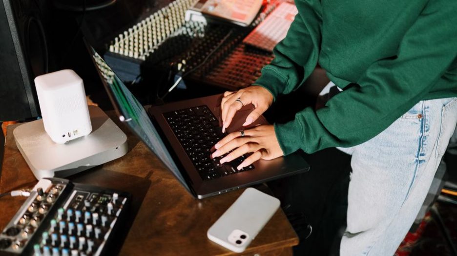 close-up of hands typing on a laptop in a recording studio surrounding audio equipment