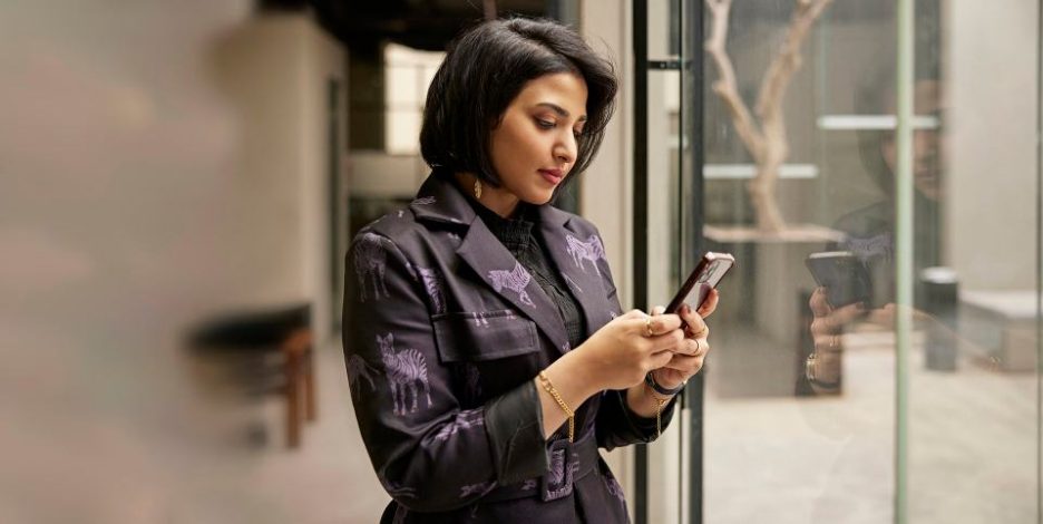 Three-quarter front view of mid 20s Saudi executive in coat-style abaya standing at window in modern office and checking mobile device.