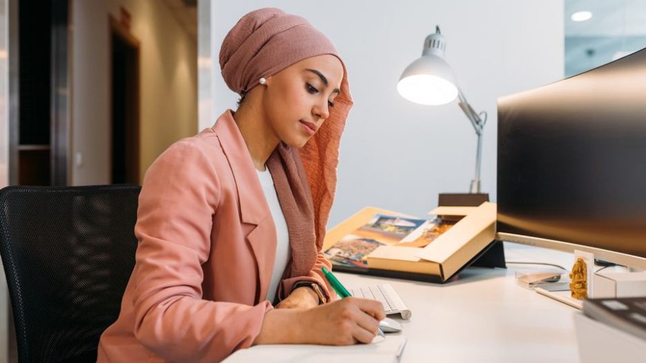 Side view of young ethnic female worker in headscarf taking notes in journal at table with computer and shiny lamp