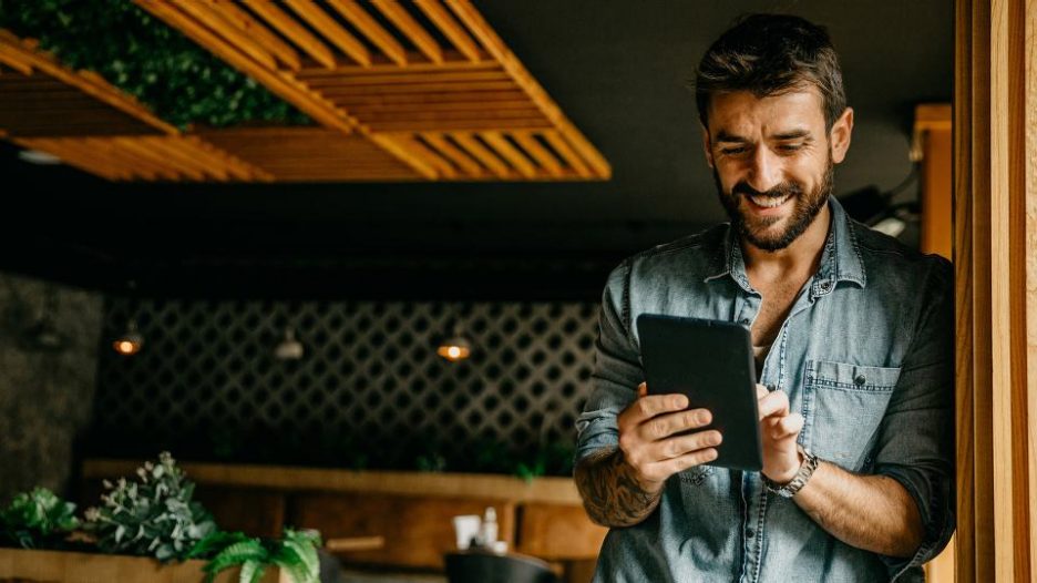 smiling man in a denim shirt is using his tablet while standing alone in a modern restaurant