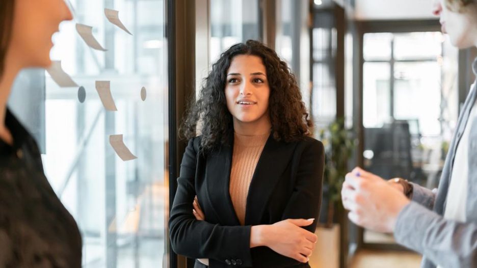 young coworkers working at modern office space near transparent collaborative board with sticky notes on it