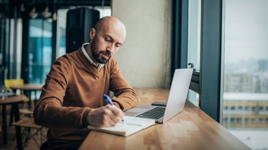 Serious businessman using laptop sitting at cafe, focused guy in suit communicating online, writing emails, distantly working or studying on computer in public place. Young businessman working on restaurant after work or in coffee break