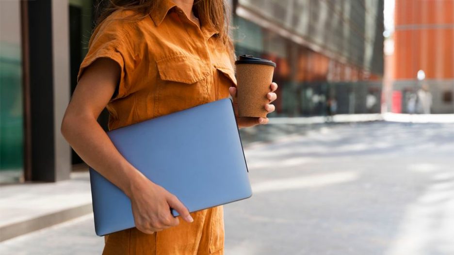 businesswoman holding laptop and takeaway coffe in front of a modern office building