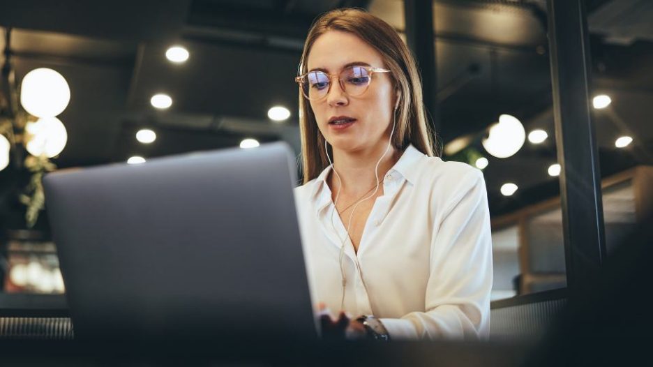 a woman in glasses is typing on her laptop while wearing earbuds in a dramatically lit office