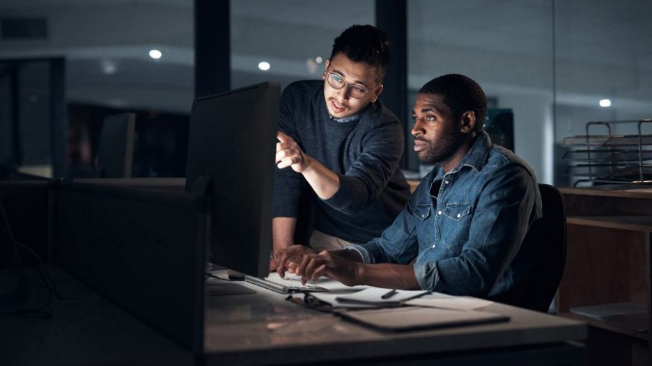 a casually dressed man is at his computer with a male coworker pointing to something on the screen while they work after hours in an office