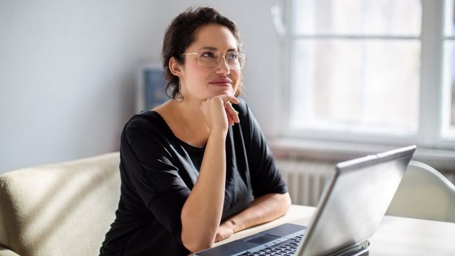 woman in a black top is thinking as she uses her laptop in an office