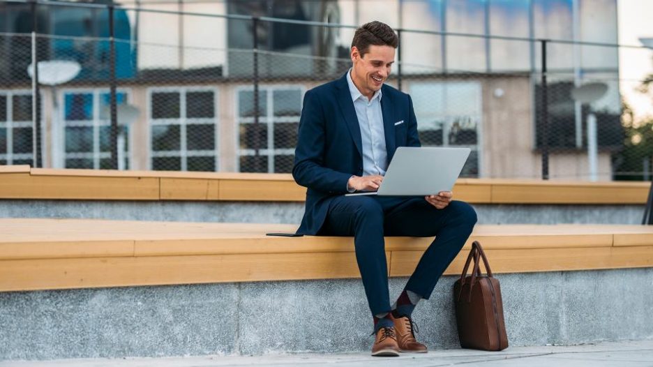 professional man with suit is sitting outside and using his latpop with offices in the background