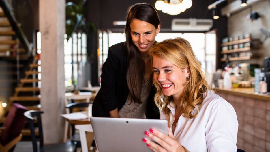 a seated women and a standing woman are smiling at a tablet while in an empty restaurant during the day