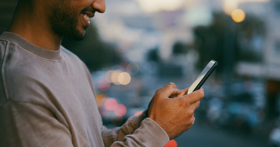 smiling man in a grey sweatshirt is using his phone on the street in the early evening