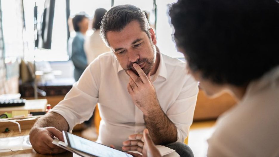 A man and woman are having a casual meetng to discuss a project at her desk and are looking at a tablet