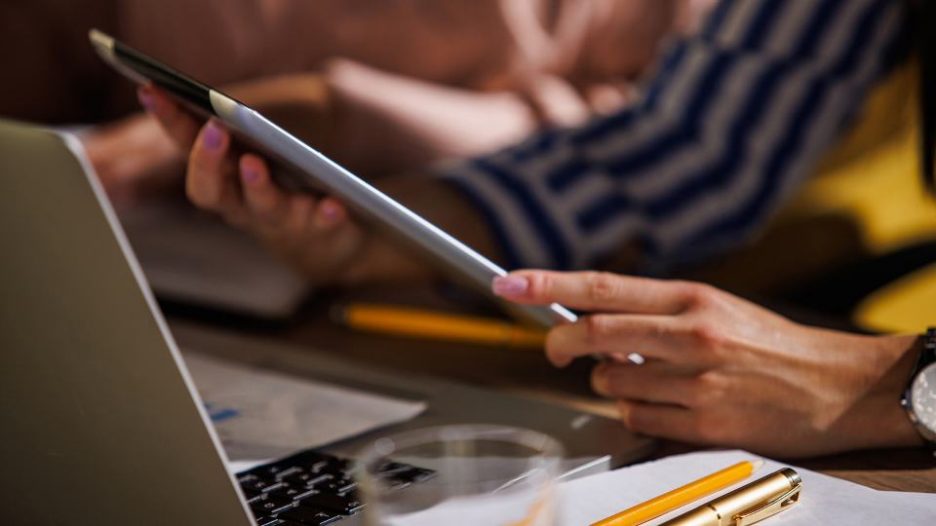 close up of a woman's hands holding a tablet with another woman in the background and a laptop nearby