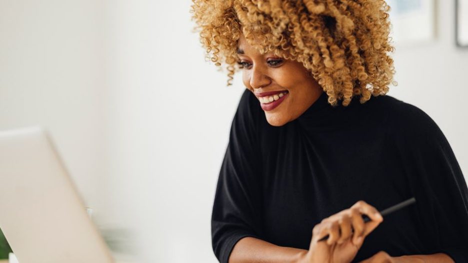 woman in black jumper against a white wall is smiling at her laptop during a conference call