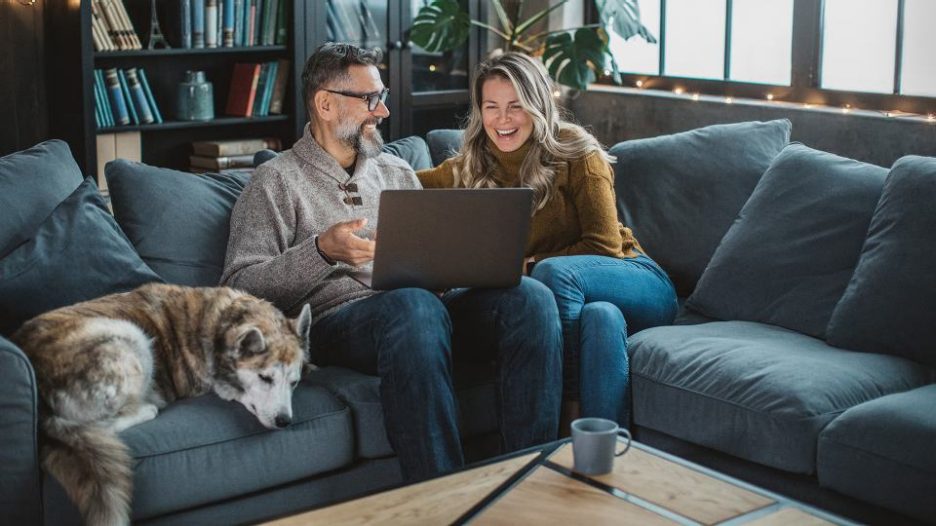 mature couple and their dog are on a blue sofa while they look at a laptop together