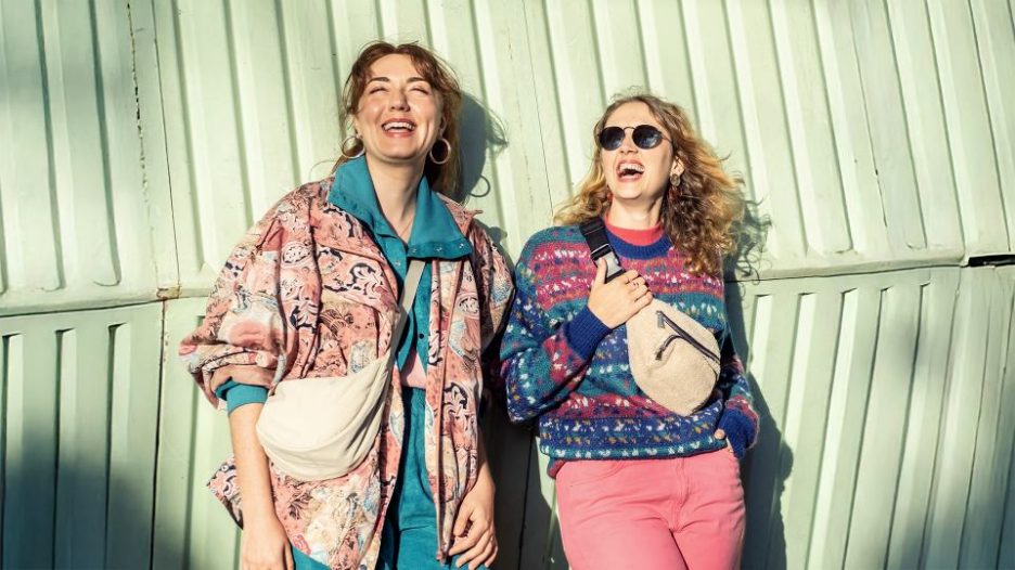 two female friends outside in front of a light green wall, laughing and enjoying life