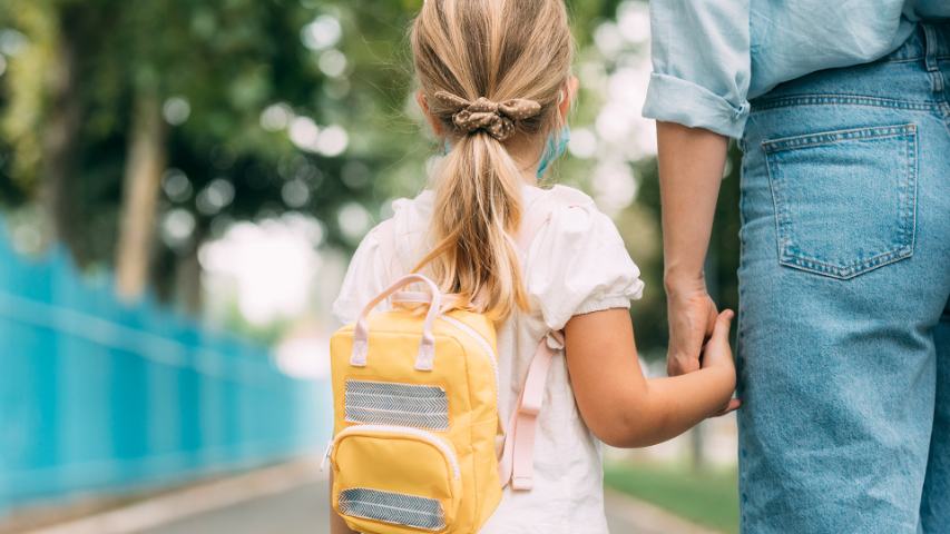 view from behind of a young girl with a back pack holding her mother's hand on the street