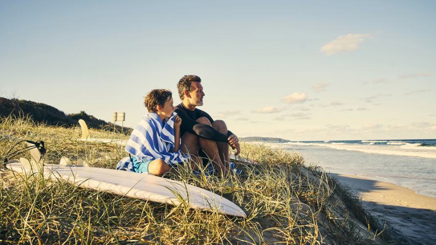 father and son sitting in the dunes with a surfboard looking out to sea