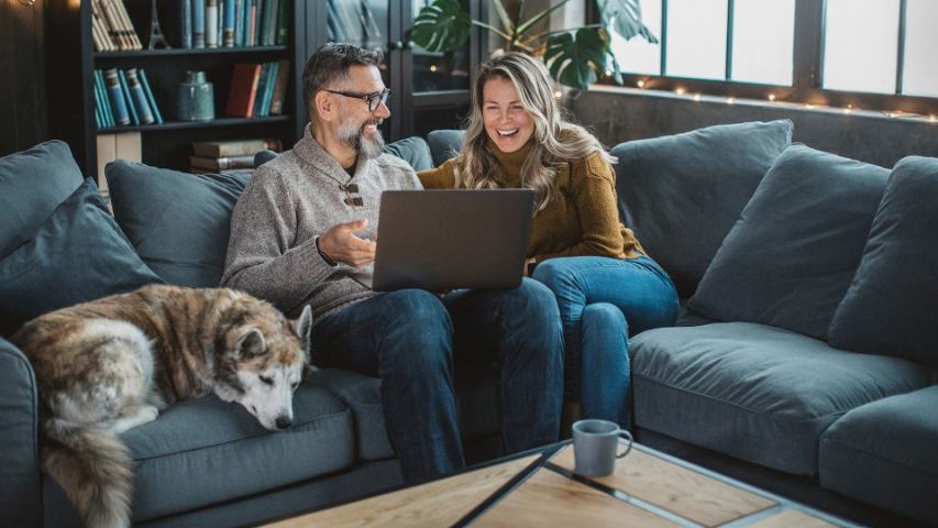 mature couple and their dog are on a blue sofa while they look at a laptop together