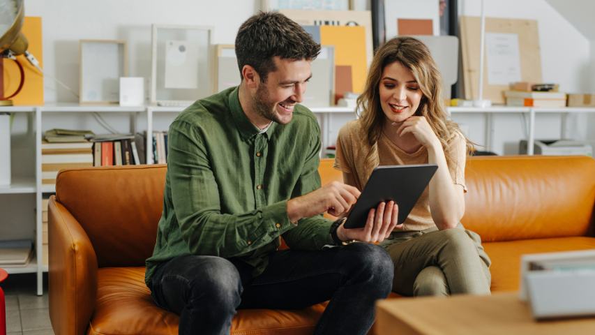 happy man and woman seated on a brown sofa looking at a tablet together