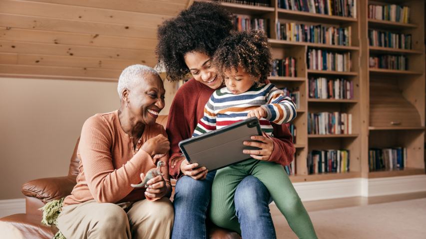 a grandmother, mother and child enjoying using a tablet while sitting on a brown sofa