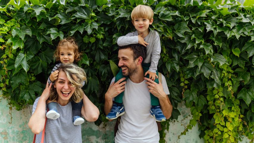 happy couple with children on their shoulders standing outside among some ivy