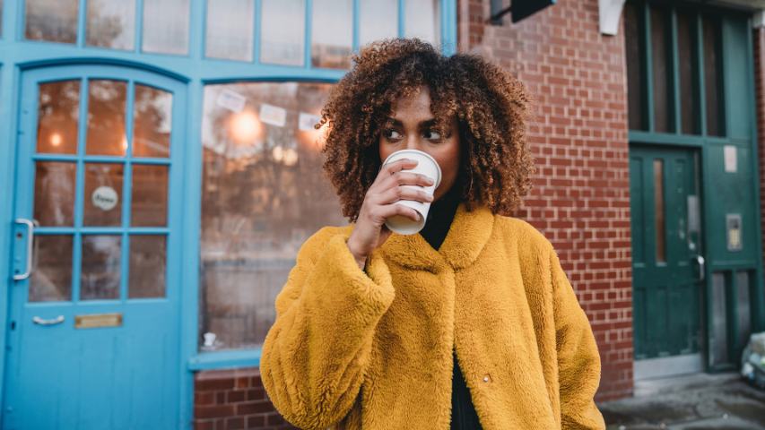 woman in a fluffy ochre coat drinking coffee outside a cafe with a blue door