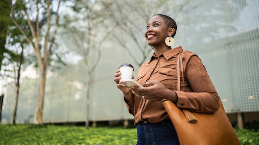 happy positive professional woman outside with her coffee, phone and handbag with an office building in the background