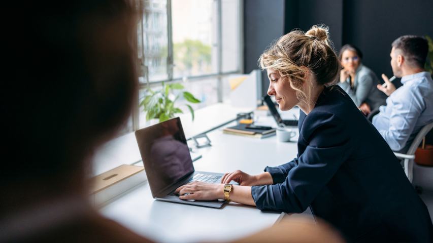 woman in a navy blazer working at a laptop in a busy modern office