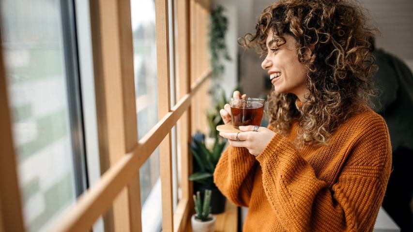 woman in a brown jumper smiling and drinking tea near a window