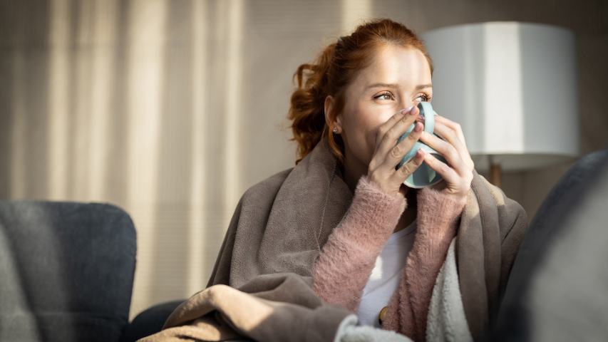 happy woman with a brown blanket and a hot drink sitting on a sofa at home