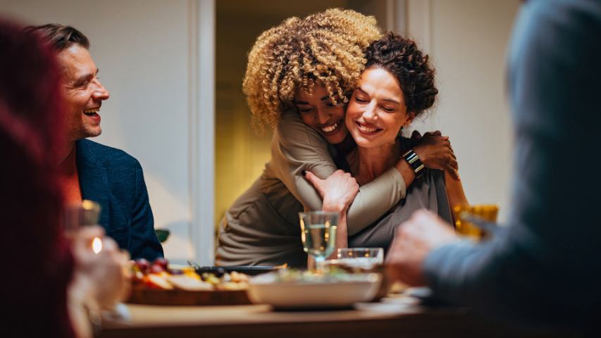 two woman hugging at a dinner party surrounded by admiring friends