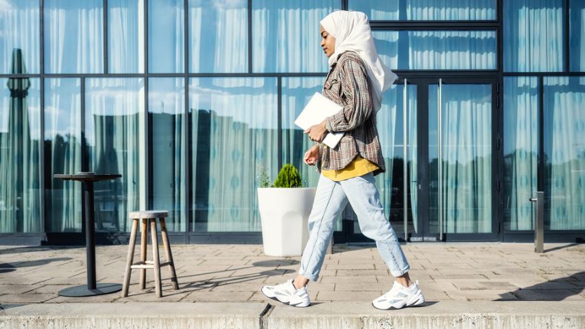 Young causualy dressed profesional woman wearing hijab and carrying laptop outside office building