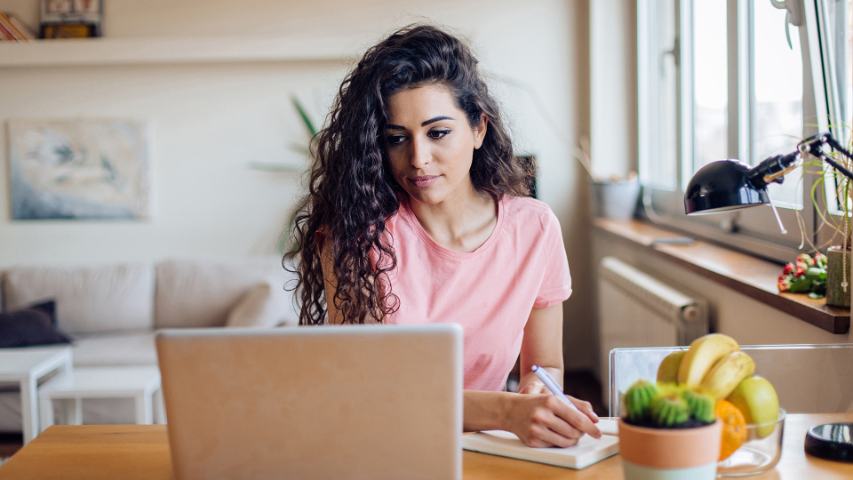 a young woman is working on her laptop at home and taking notes