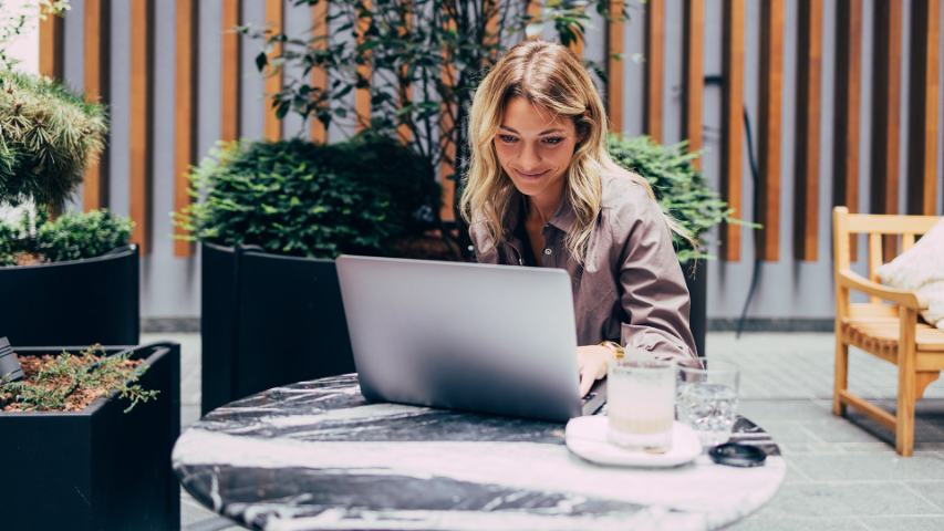 a woman working at a cafe is seated outdoors and smiling at her laptop