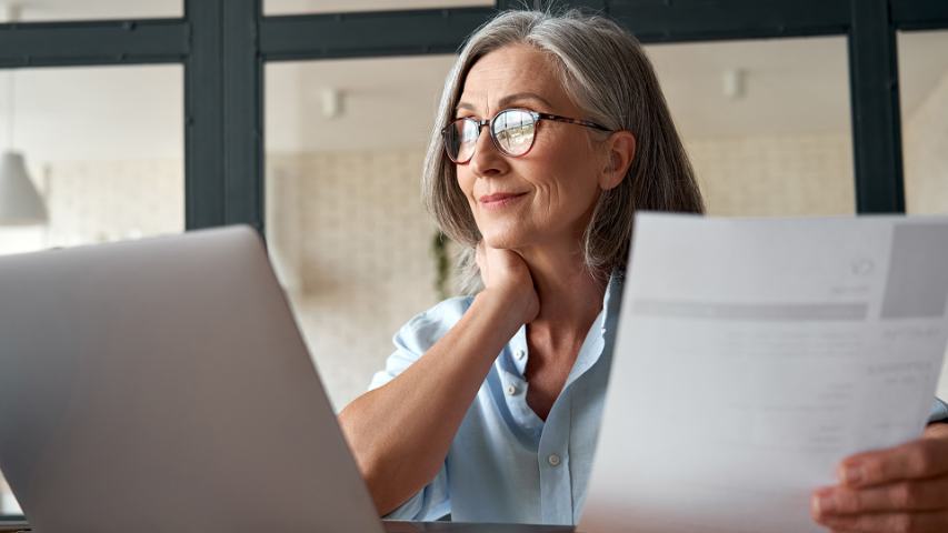 woman with glasses pauses thoughtfully while using a laptop and reviewing paperwork