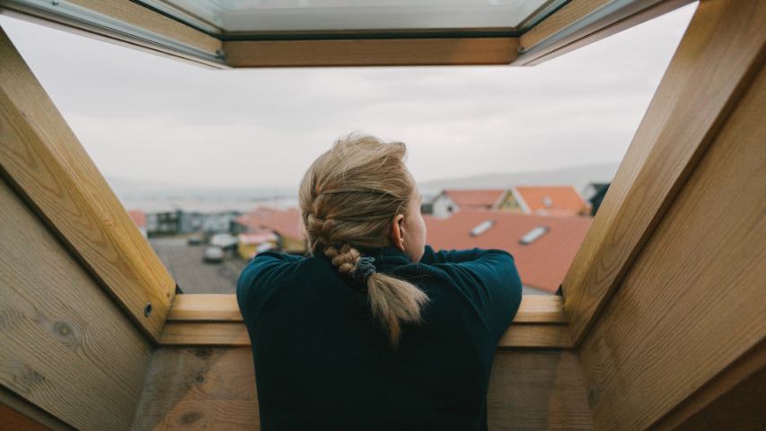 view from behind of a young woman looking out a timber framed window to the rooftops beyond