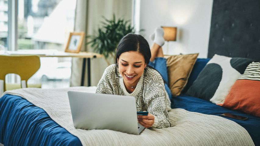 young woman lying on her bed and using her laptop and credit card