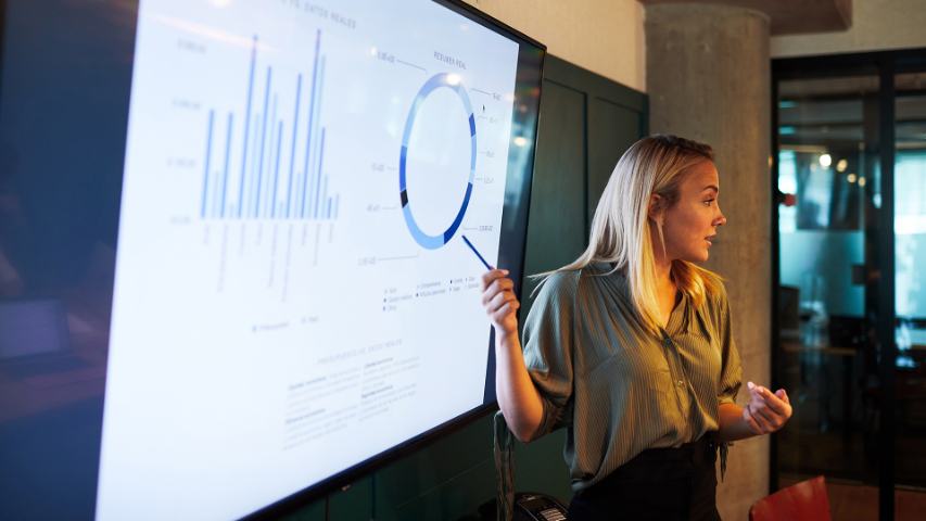 woman standing at a large monitor and pointing to a graph while making her presentation