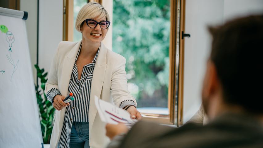 smiling woman in glasses and a blazer is presenting using a whiteboard and is handing papers to a male oworker