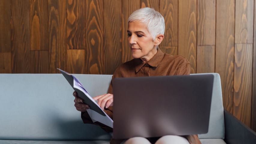 Female professional sitting on office sofa while reading notes and balancing laptop on her lap