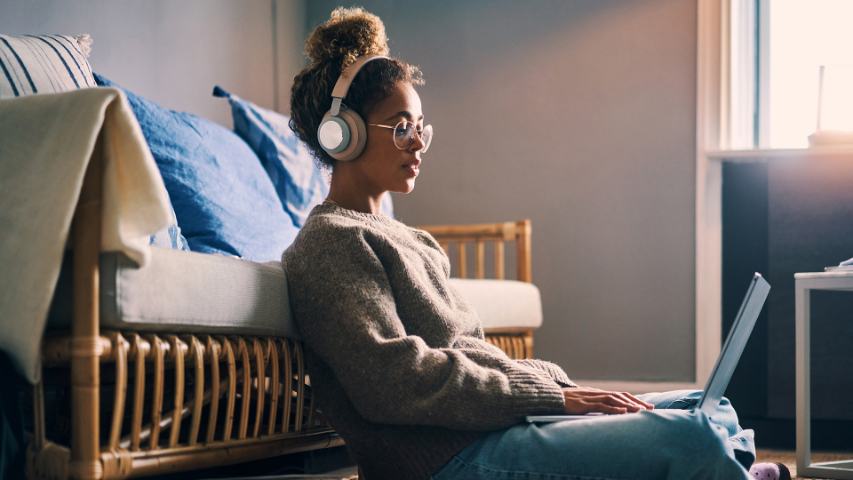 young woman with glasses sitting up against a sofa with her headphones and laptop