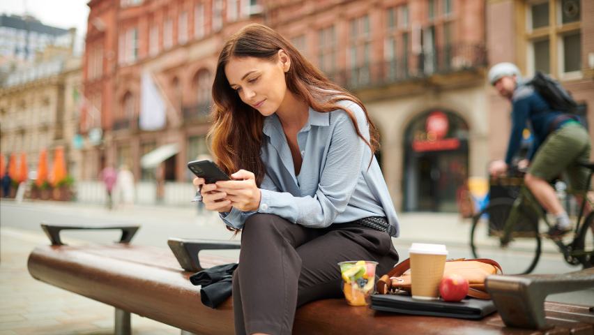 woman sitting on a bench outside, using her phone with her lunch and coffee nearby